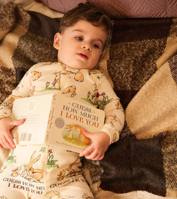 Baby wearing bunny print coverall holding "Guess How Much I Love You" book